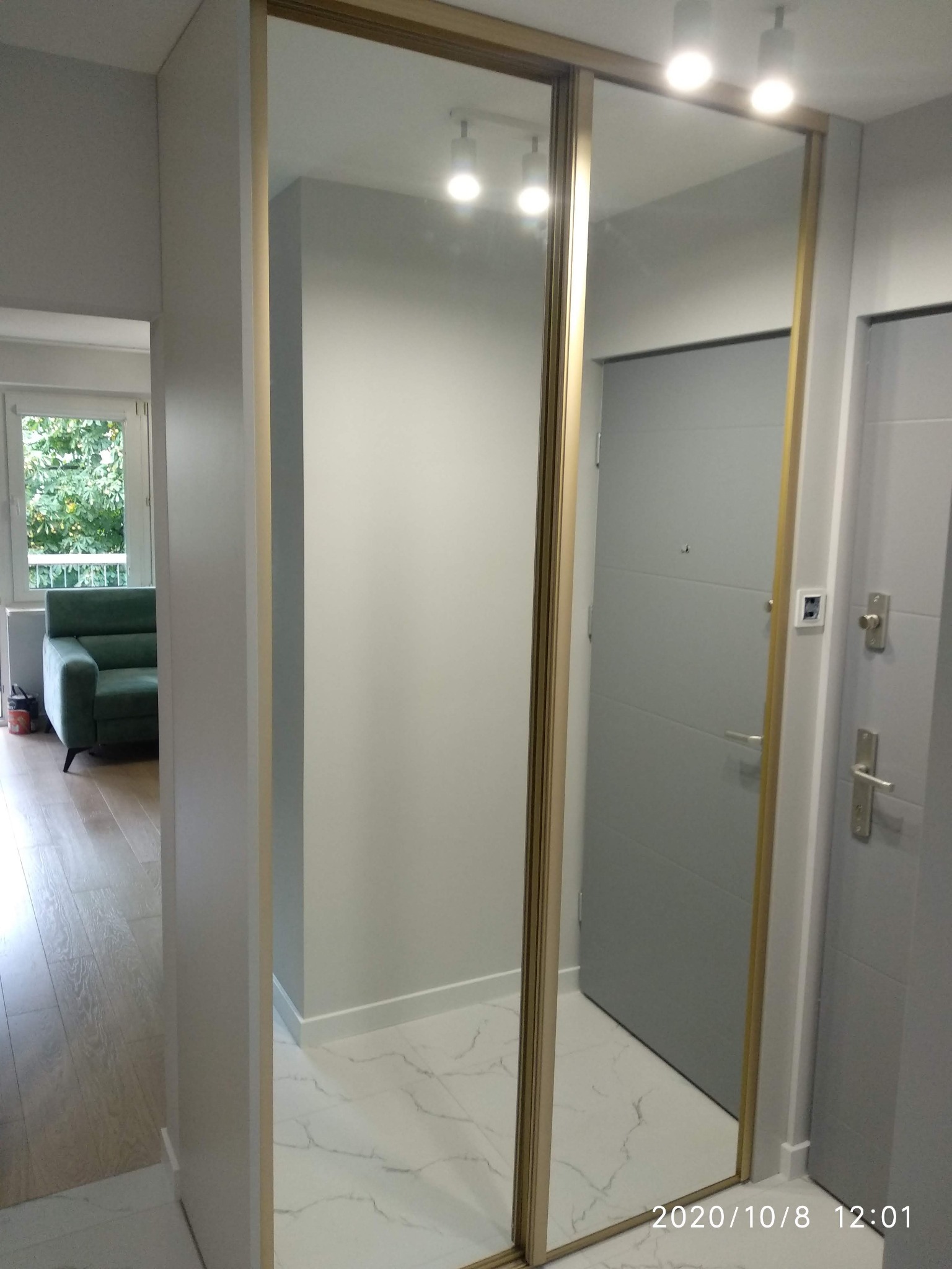 Nowoczesna szafa z przesuwnymi drzwiami lustrzanymi, złote obramowanie, widok na korytarz z zielonym fotelem i drzwi wejściowe.