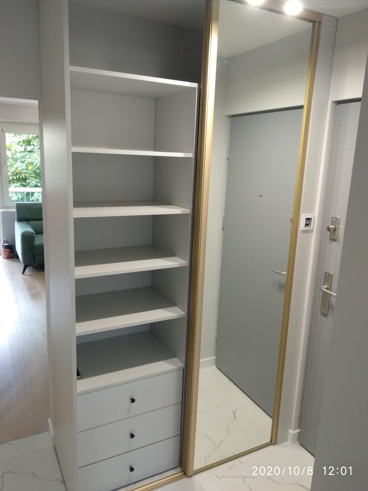 Biała szafa z półkami i trzema szufladami, z lustrzanymi drzwiami przesuwnymi, odbijająca szarą ścianę i drzwi wejściowe, widoczna z perspektywy wnętrza mieszkania.