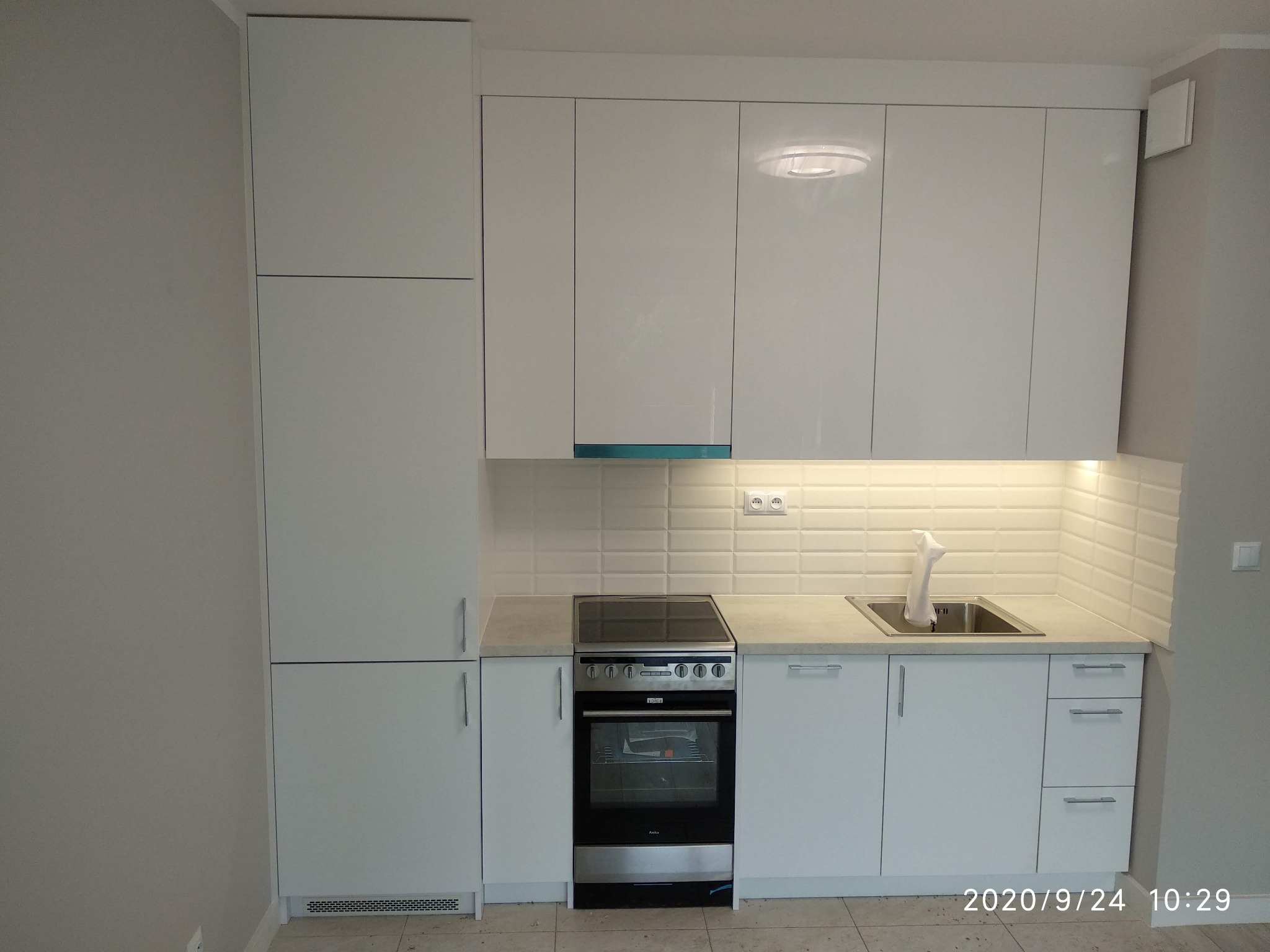 Biała zabudowa kuchenna z płytkami imitującymi cegłę na ścianie, widoczny zlewozmywak, płyta grzewcza i piekarnik Amica.