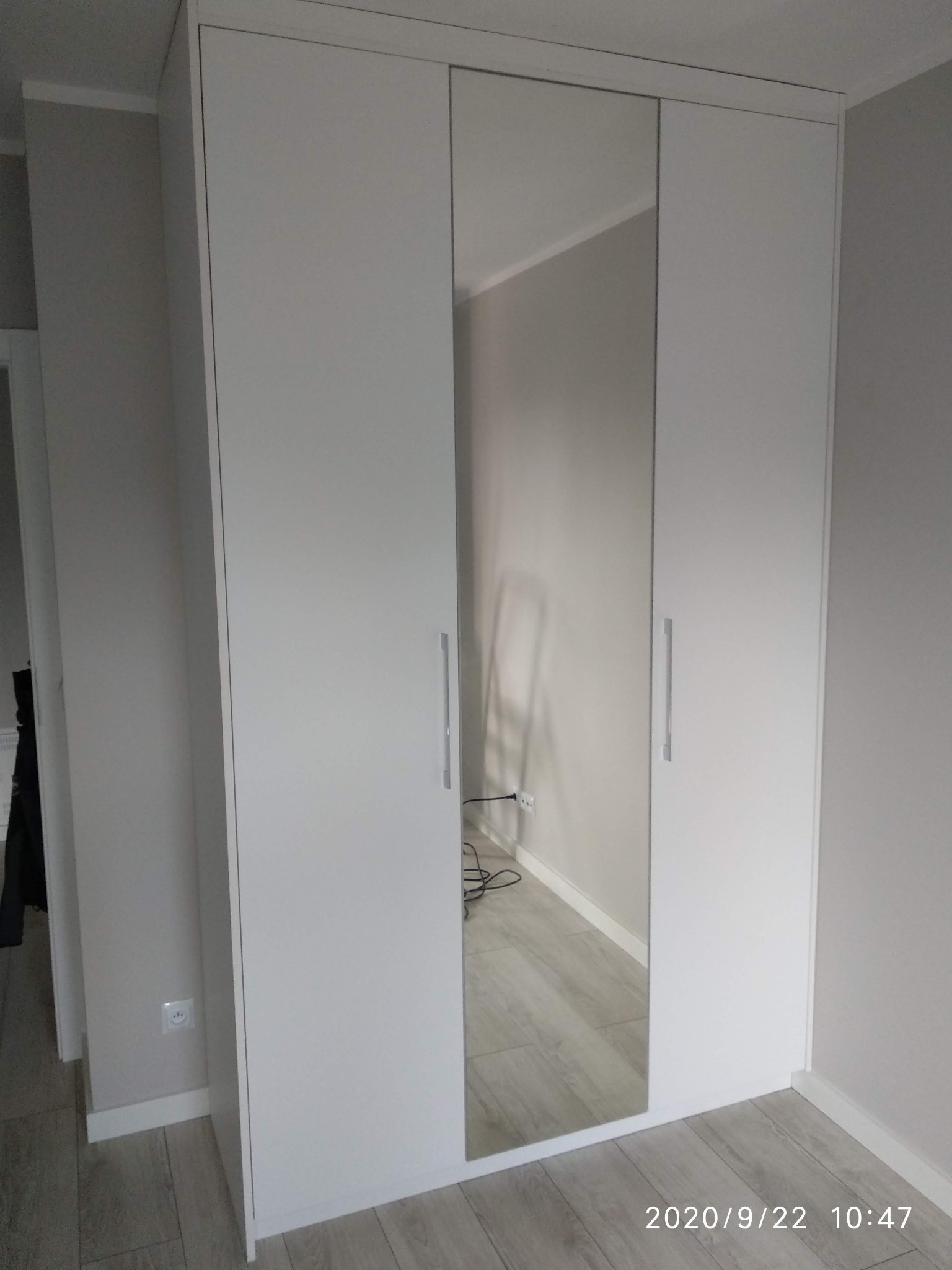 Biała szafa wnękowa z przesuwnymi drzwiami, jedno skrzydło z lustrem odbija wnętrze pokoju z szarą podłogą i ścianami.