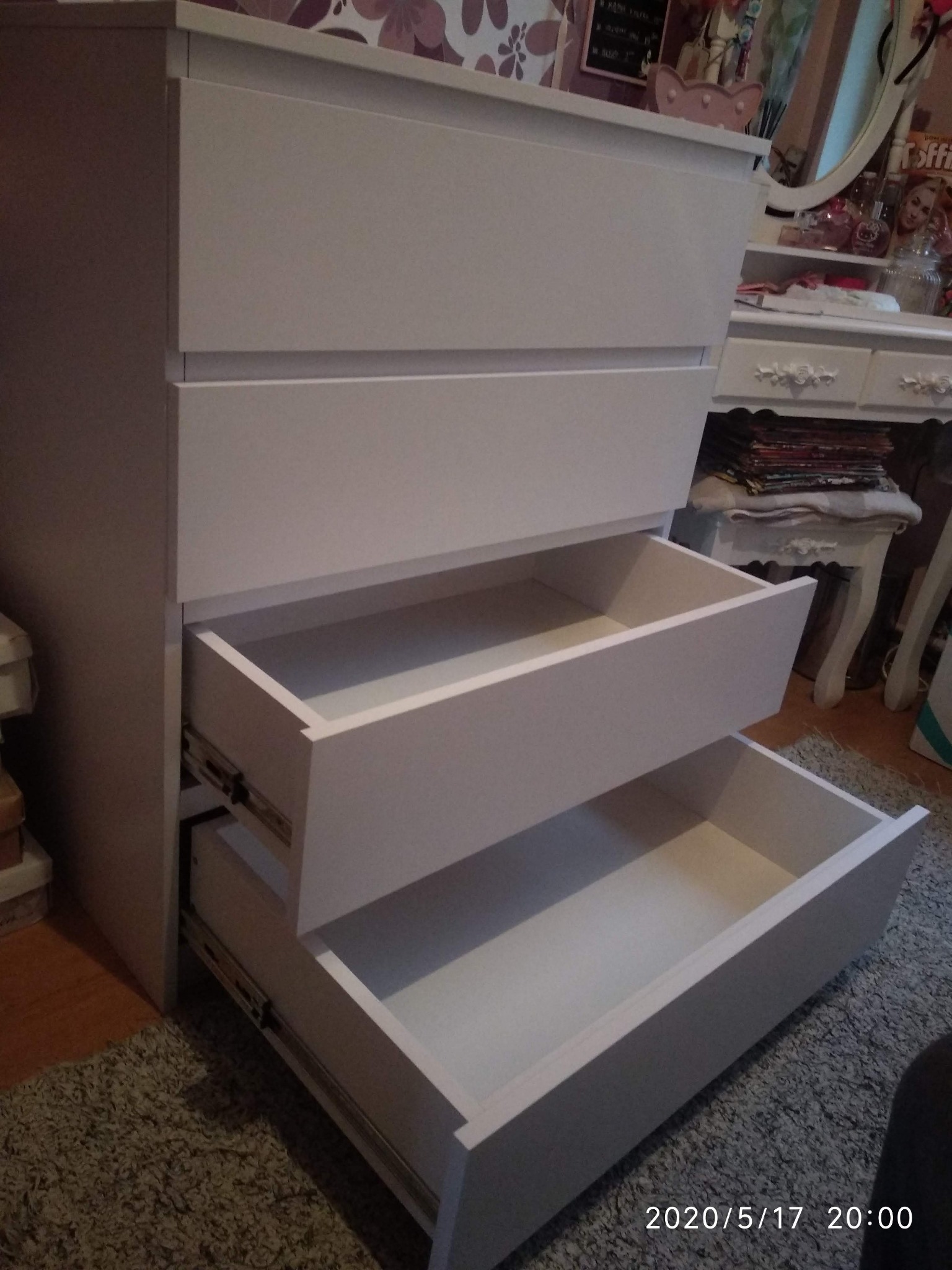 Biała komoda z czterema szufladami, dwie górne otwarte, ukazujące puste wnętrze, widoczne prowadnice szuflad, w tle toaletka z lustrem i akcesoriami.