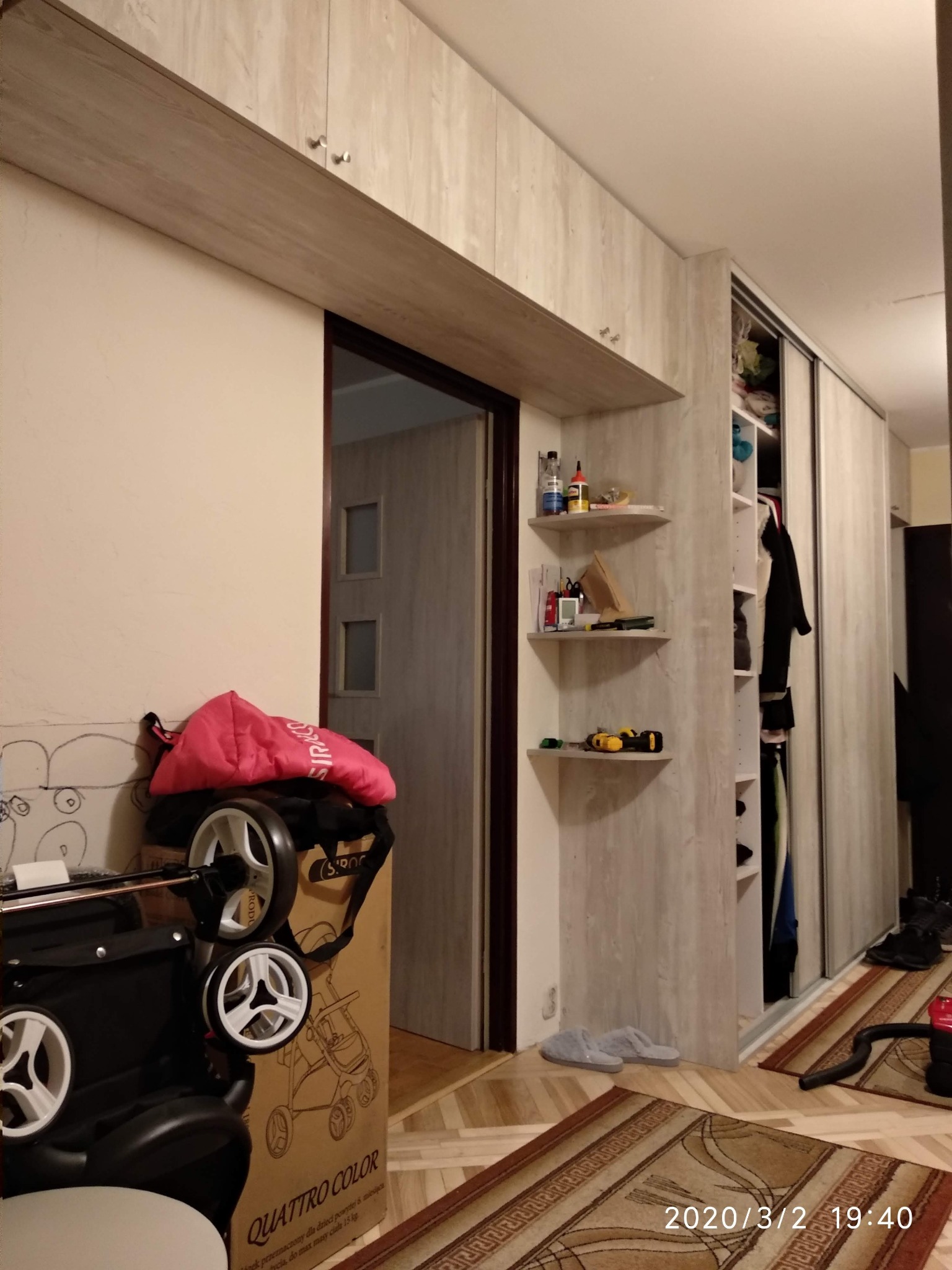 Wnętrze pokoju z zabudową meblową w odcieniach beżu i szarości, obejmującą szafę przesuwną i półki narożne, z widocznym w tle wejściem do innego pomieszczenia. Na pierwszym planie karton po wózku...