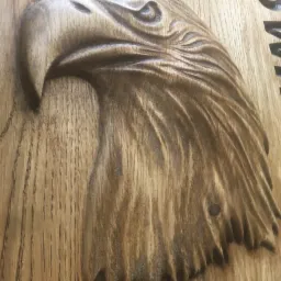 Ręcznie rzeźbiona głowa orła w drewnie z widocznymi słojami, fragment napisu w tle.