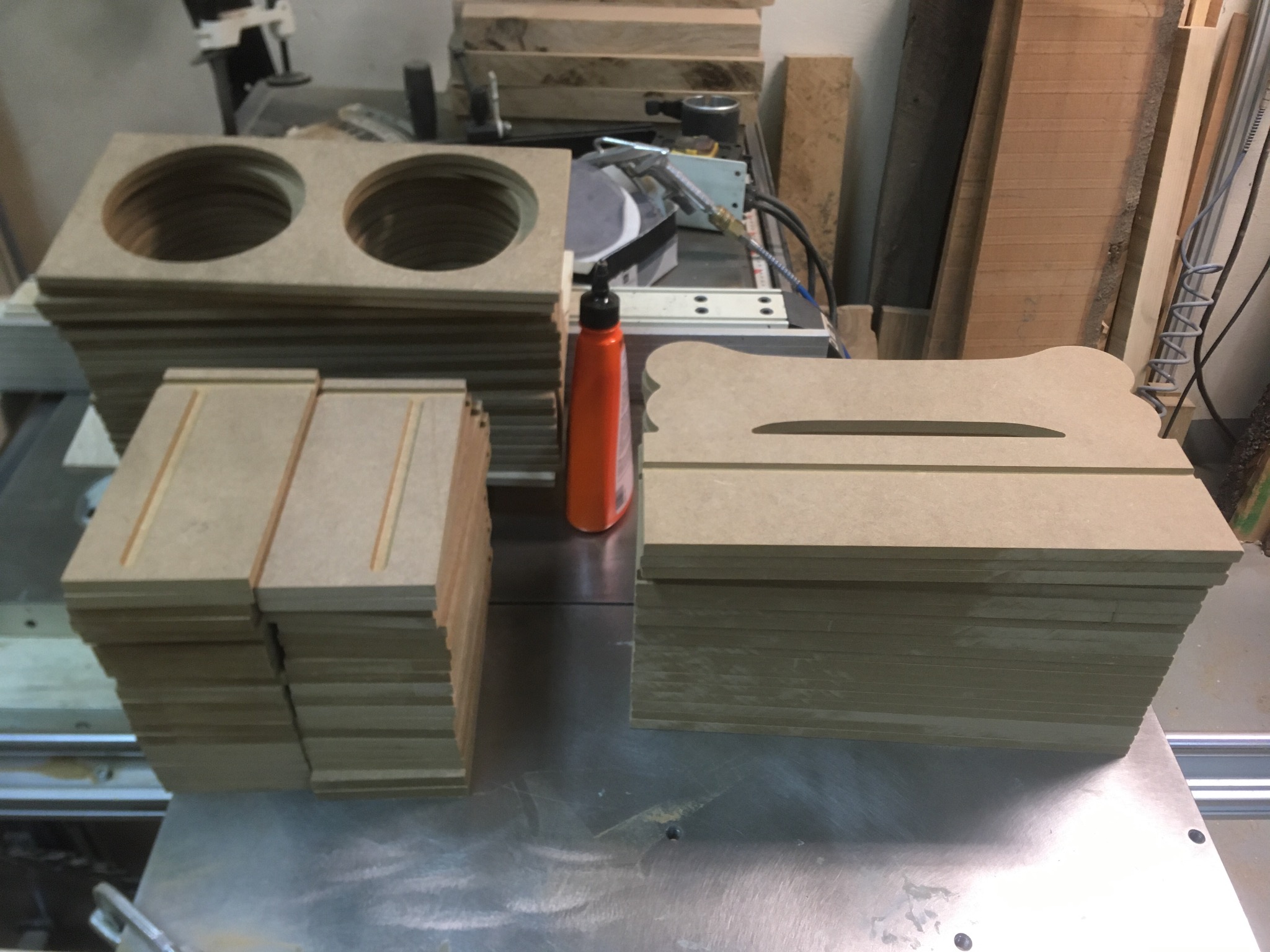 Stosy wyciętych elementów z płyty MDF o różnych kształtach na metalowym stole warsztatowym; widoczne otwory, szczeliny i krawędzie, w tle narzędzia i materiały.