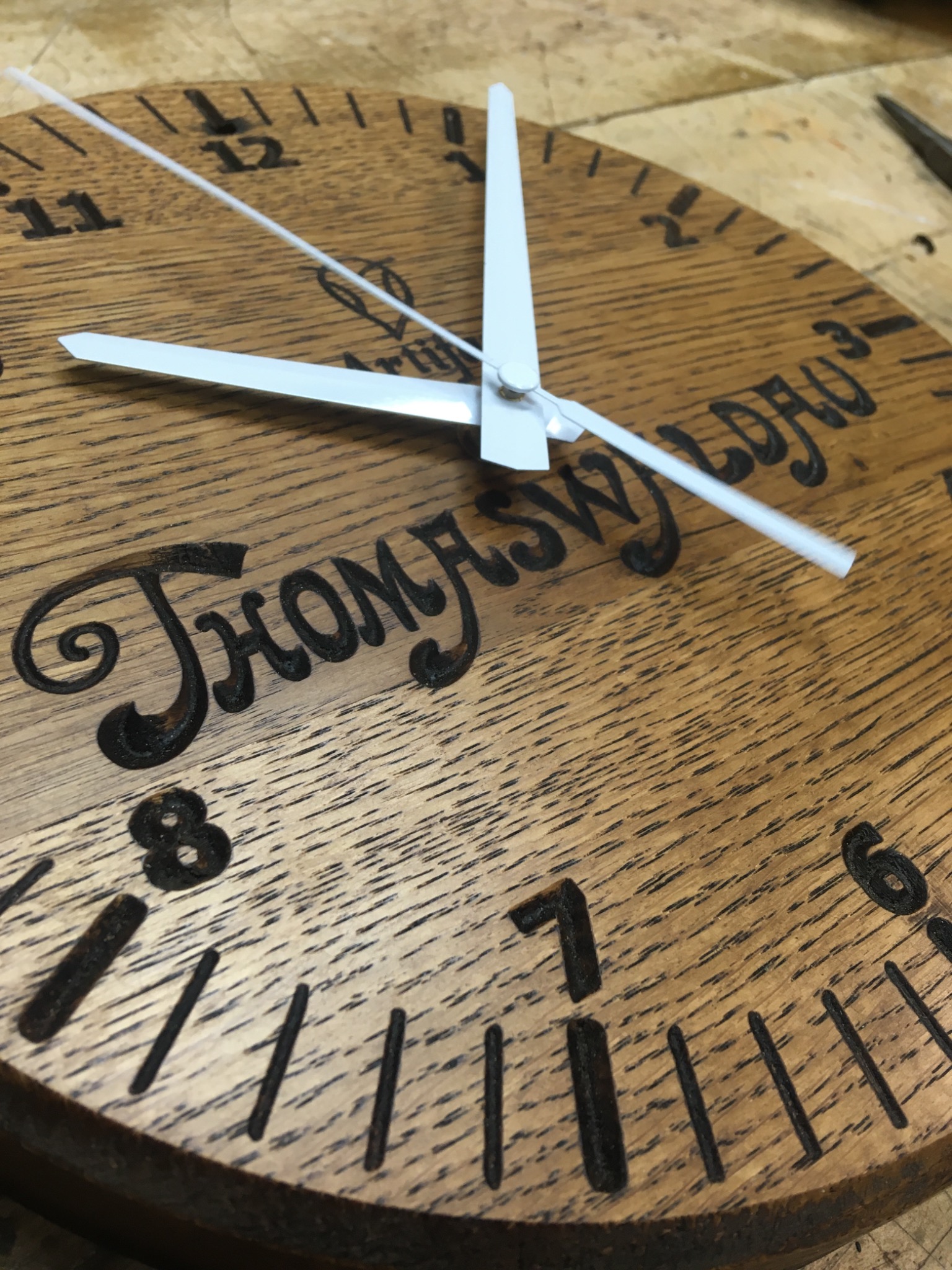Drewniany zegar z wygrawerowanym napisem 'Thomysw Lordu' i białymi wskazówkami, widoczne detale faktury drewna i wyżłobione cyfry.