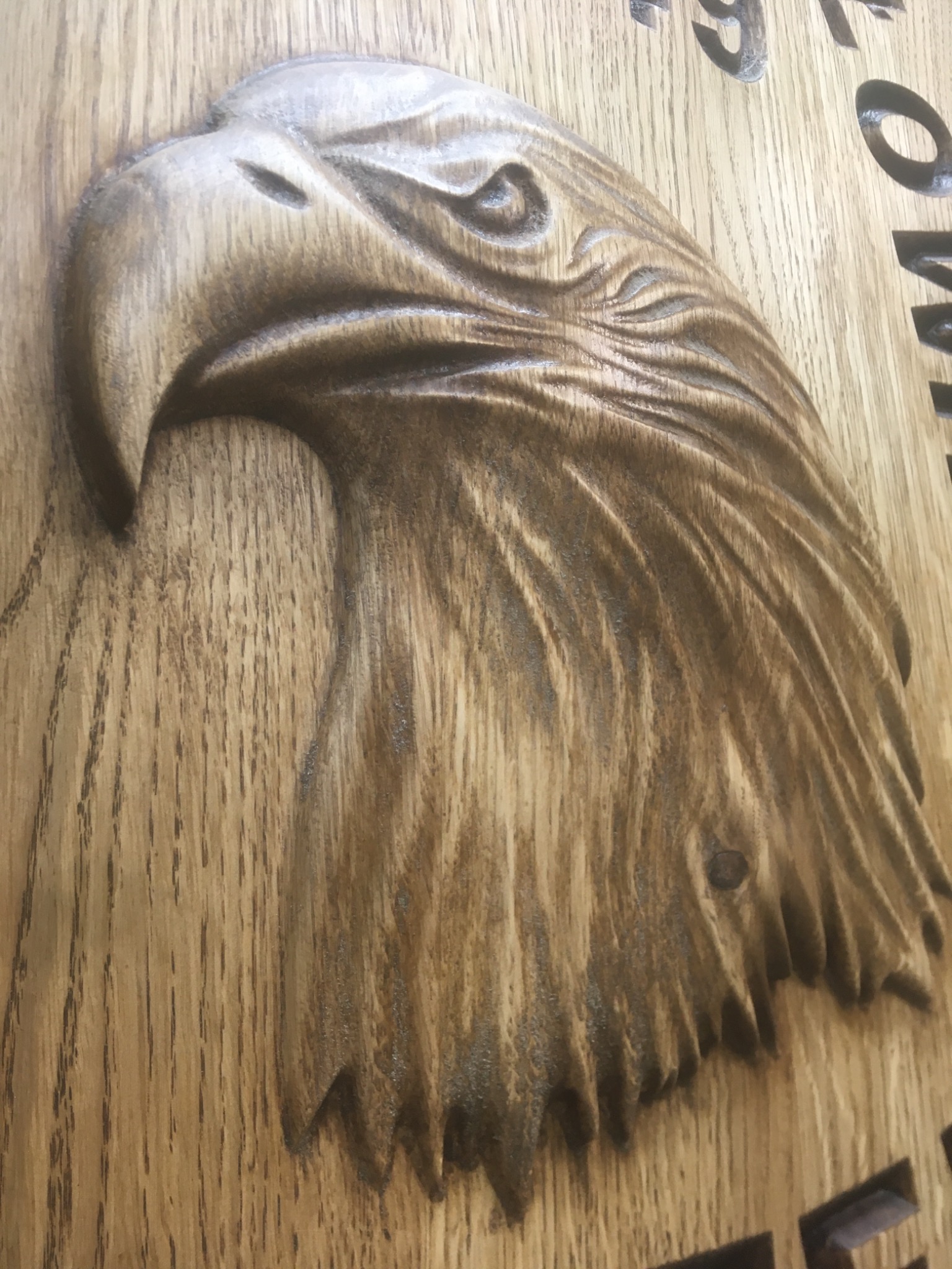 Ręcznie rzeźbiona głowa orła w drewnie z widocznymi słojami, fragment napisu w tle.