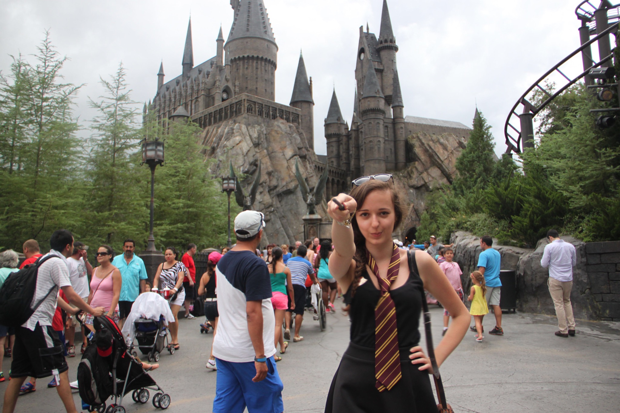 Kobieta w czarnej sukience i krawacie w paski, trzymająca różdżkę na tle zamku Hogwart i tłumu ludzi w parku rozrywki.