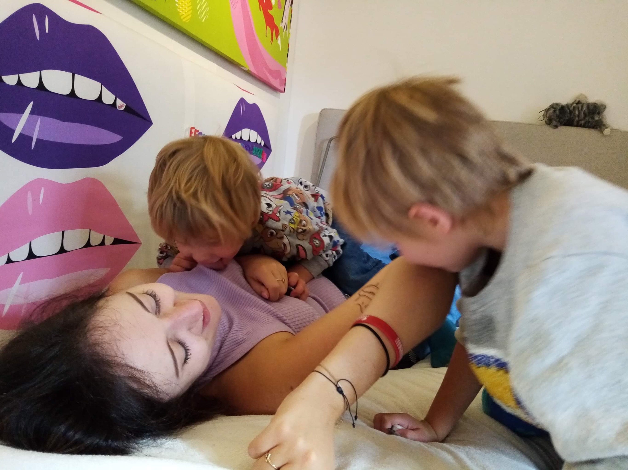 Kobieta leżąca na łóżku, przytulana przez dwóch chłopców, z kolorowymi grafikami ust na ścianie w tle.