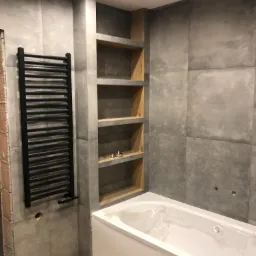 Nowoczesna łazienka w szarościach z drewnianymi półkami wnękowymi, czarnym grzejnikiem drabinkowym i białą wanną, jeszcze zabezpieczoną folią ochronną.