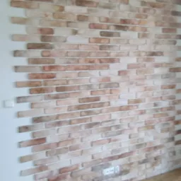 Ściana wykończona dekoracyjną cegłą w odcieniach beżu i brązu, ułożona poziomo z widoczną fugą, fragment białego sufitu i ściany, widoczny kontakt elektryczny.