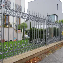 Szare, metalowe ogrodzenie z dekoracyjnymi elementami roślinnymi i geometrycznymi, oddzielające posesję z budynkiem mieszkalnym od chodnika.
