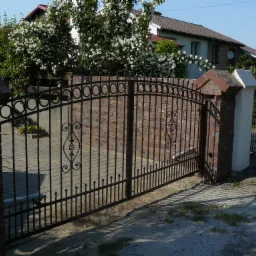 Kuta brama wjazdowa z ceglanymi słupkami, widoczny automat do bramy, tło z kwitnącymi krzewami i domami.