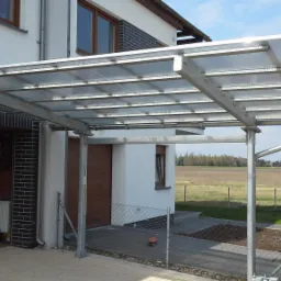 Zadaszenie garażowe z metalową konstrukcją i przezroczystym pokryciem, widoczne opony w garażu, drabina i grzejnik, dom jednorodzinny w tle.