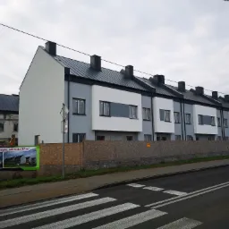 Nowoczesne szeregowe domy w trakcie budowy, z szarą i białą elewacją, czarnym dachem, widoczne zabezpieczenia okien folią oraz budowa kolejnego segmentu w tle.