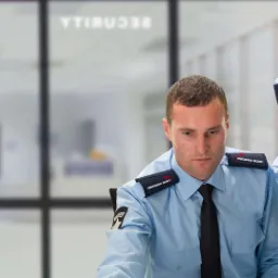 Dwóch ochroniarzy w błękitnych uniformach 'Professional Security', kobieta używa krótkofalówki, mężczyzna patrzy w dół, w tle przeszklone biuro z napisem 'SECURITY'.