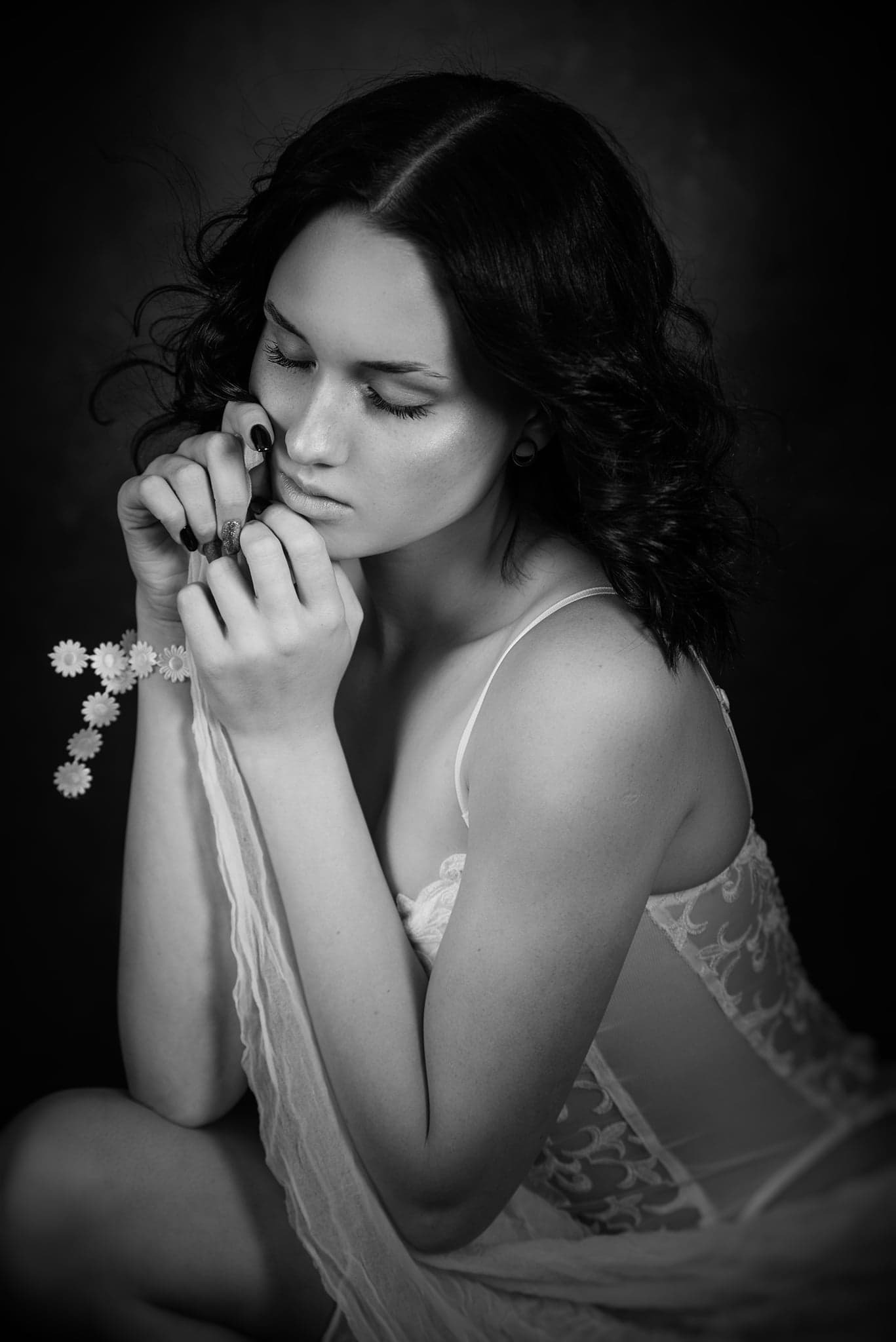 Portret młodej kobiety w bieliźnie z koronką, z zamkniętymi oczami, trzymającej w dłoniach wianek z białych kwiatów na ciemnym tle, w czerni i bieli.
