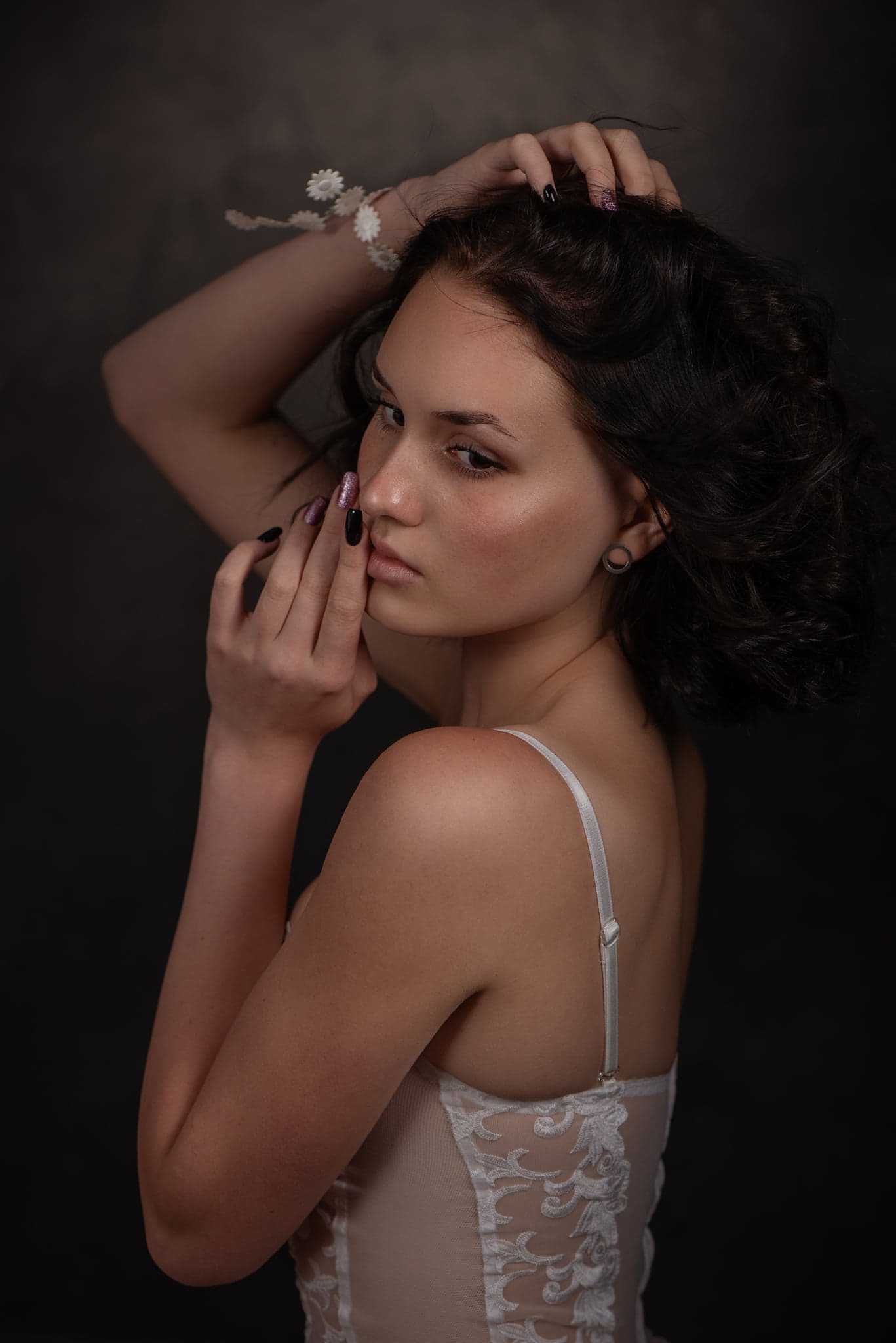 Portret młodej kobiety z ciemnymi włosami, w jasnej bieliźnie, z delikatnym makijażem, pozującej na ciemnym tle w studio. Dłonie przy twarzy, subtelna ozdoba na włosach.