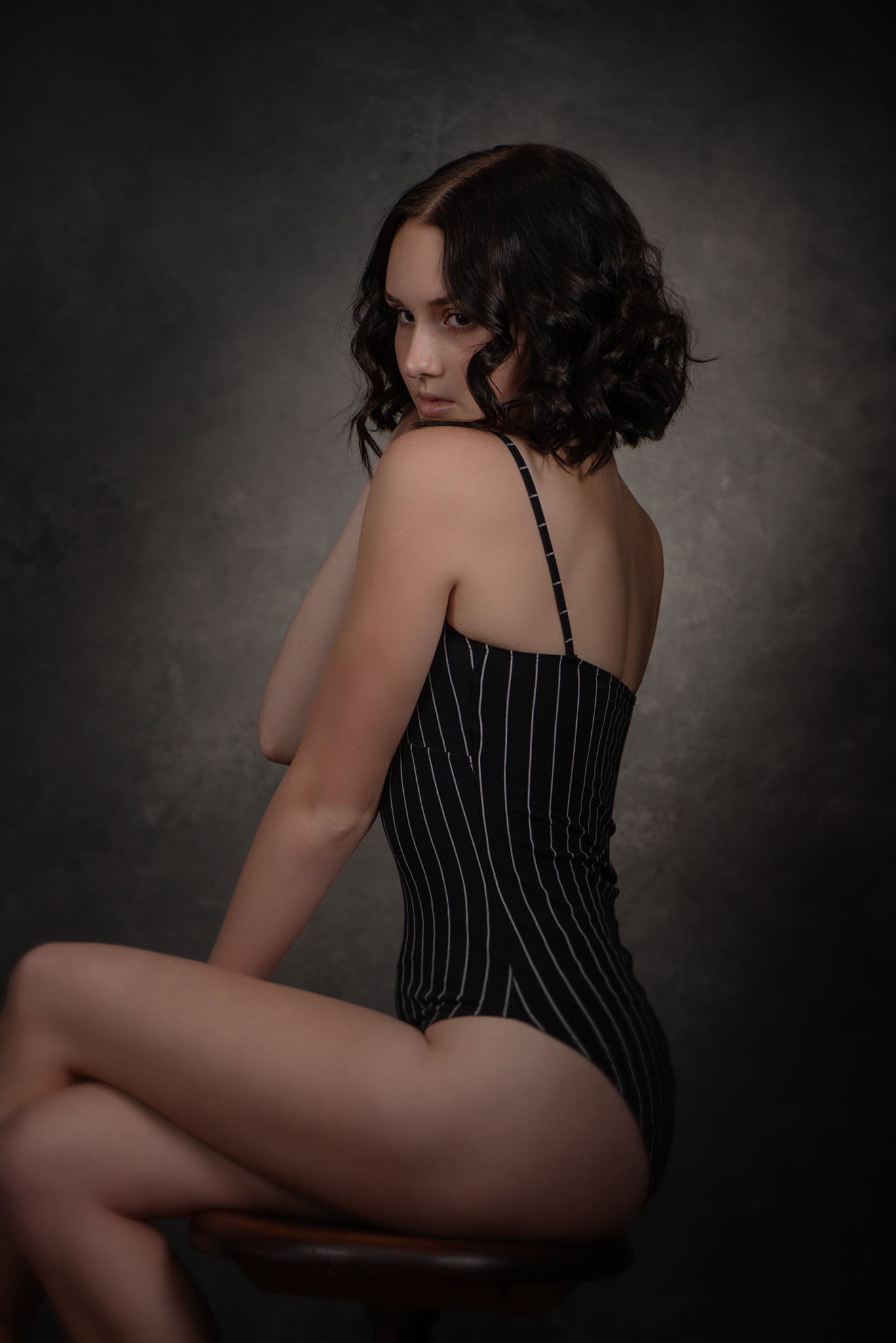 Portret kobiety w czarno-białym body w paski, siedzącej na drewnianym stołku na tle szarego tła, z głową odwróconą w stronę widza.