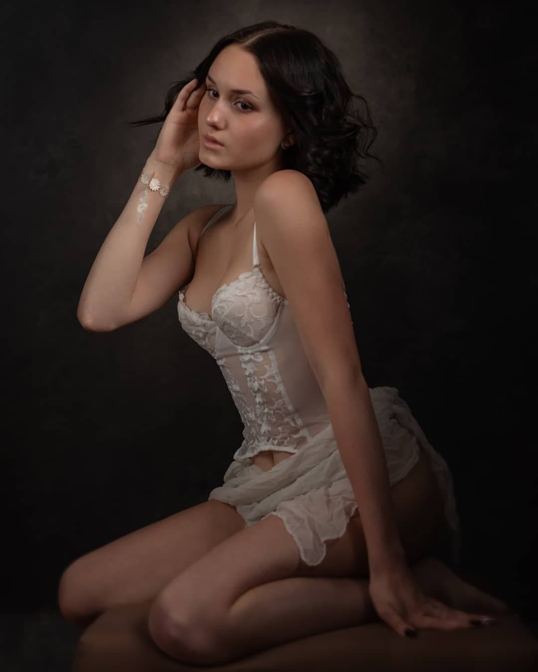 Portret młodej kobiety w białej bieliźnie, siedzącej na kolanach na ciemnym tle, z delikatnym bransoletką z motywem kwiatowym na ramieniu.