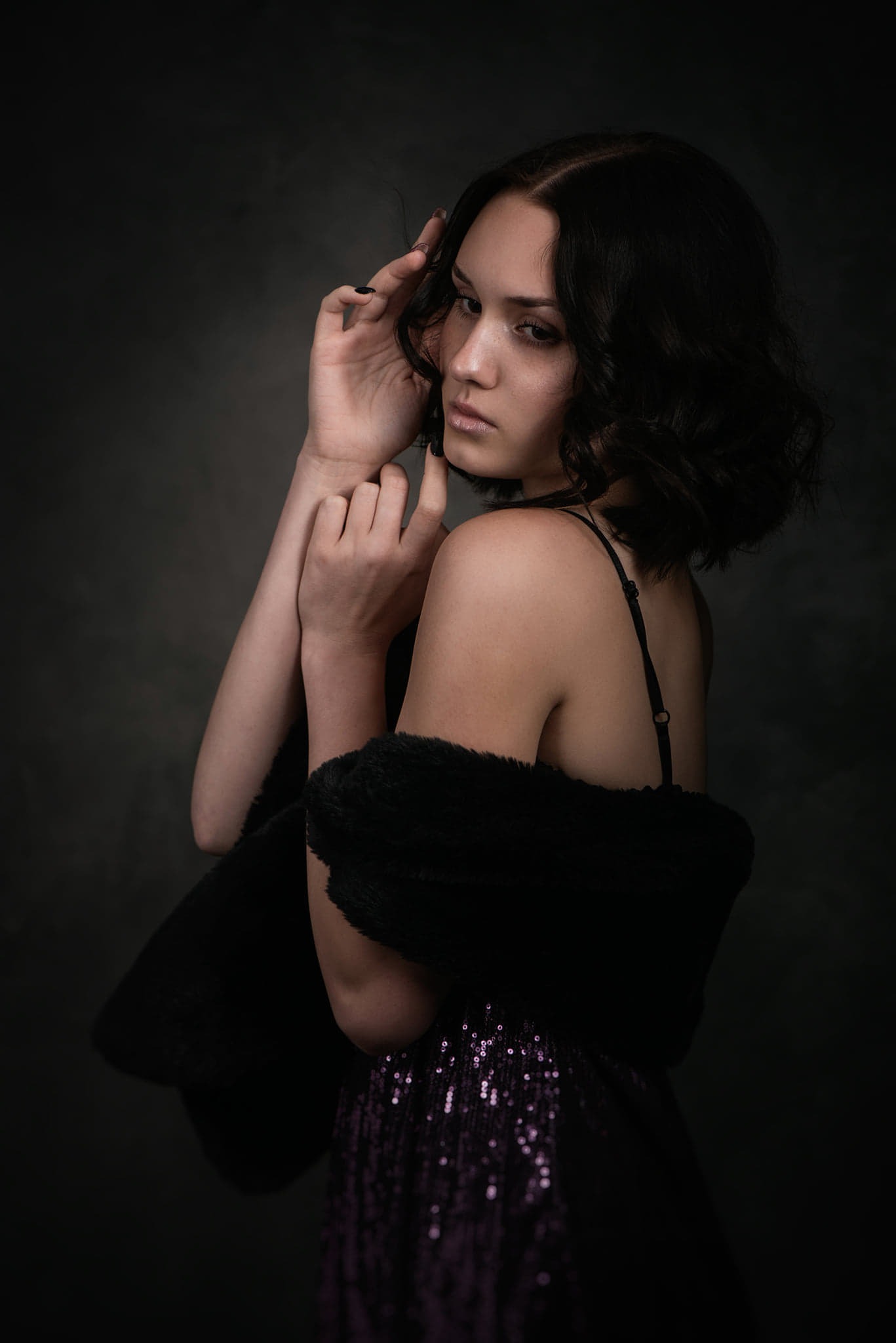 Portret młodej kobiety w ciemnej sukience z cekinami i czarnym futrzanym szalem, pozującej na ciemnoszarym tle. Delikatne oświetlenie podkreśla jej rysy twarzy i elegancki wygląd.