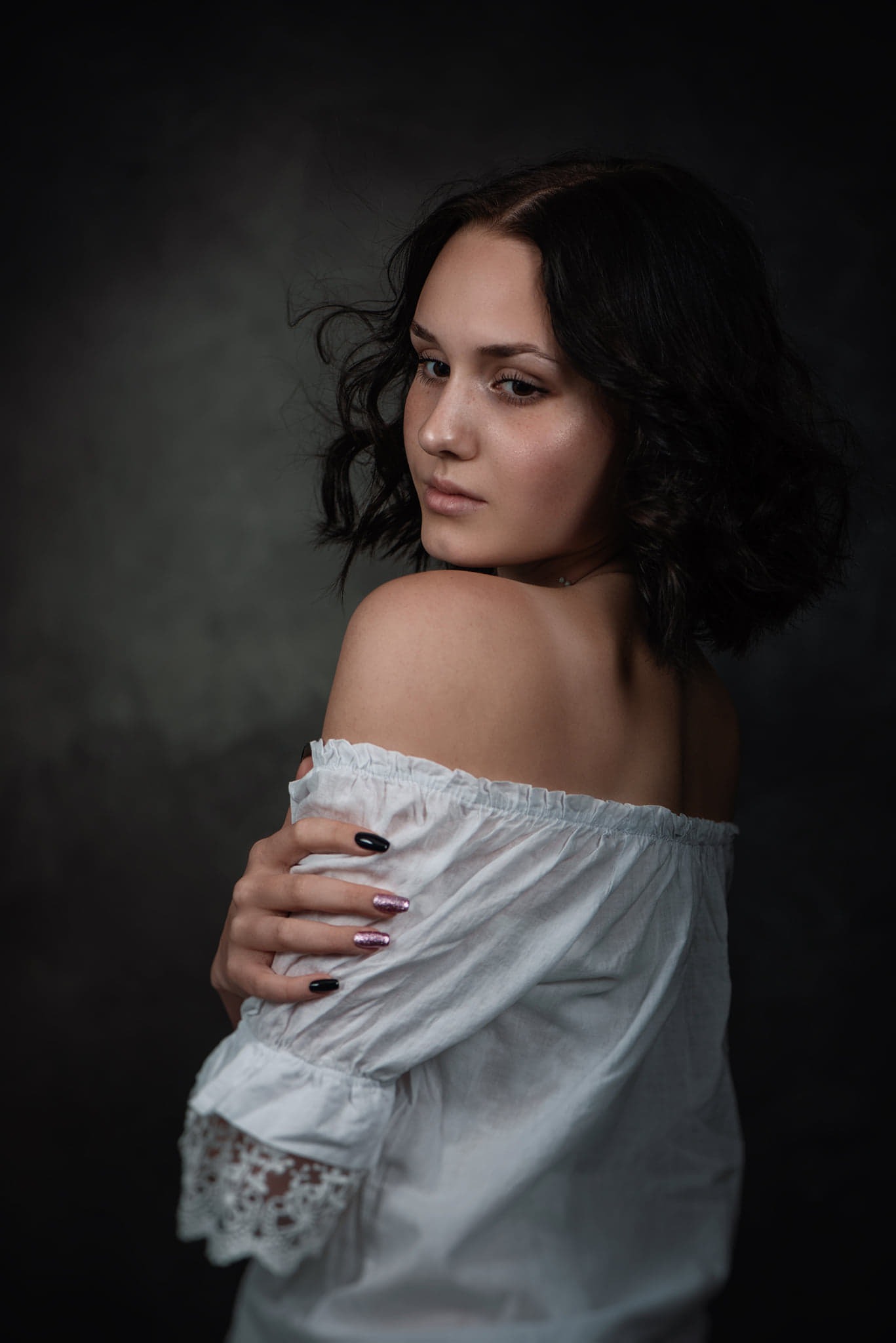 Portret młodej kobiety z ciemnymi kręconymi włosami, odwróconej przez ramię, w białej bluzce z odkrytymi ramionami i koronkowymi mankietami na tle ciemnego, rozmytym tle.