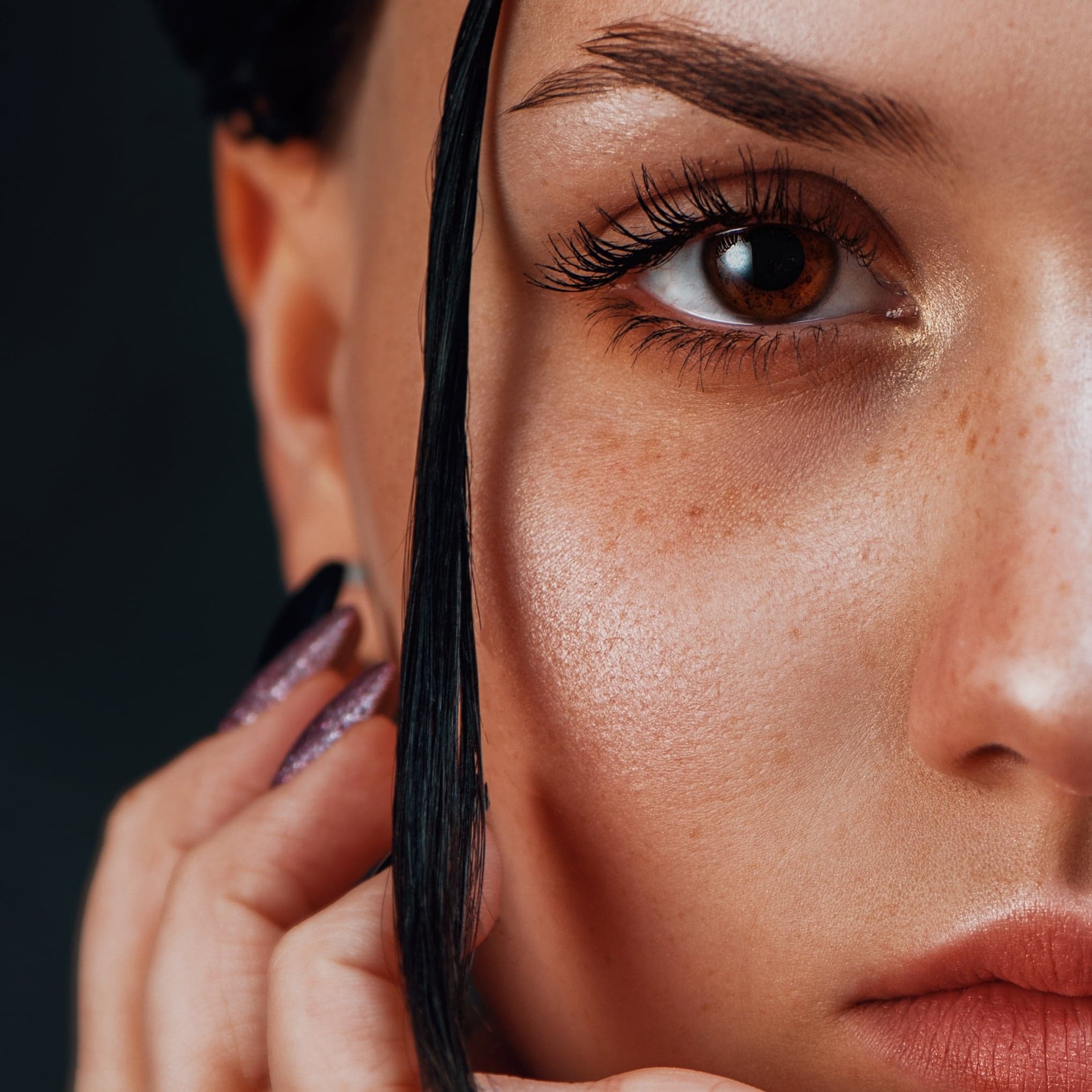 Portret zbliżony twarzy kobiety z brązowym okiem, piegami i długimi rzęsami, czarne włosy opadają na twarz, błyszczące fioletowe paznokcie.