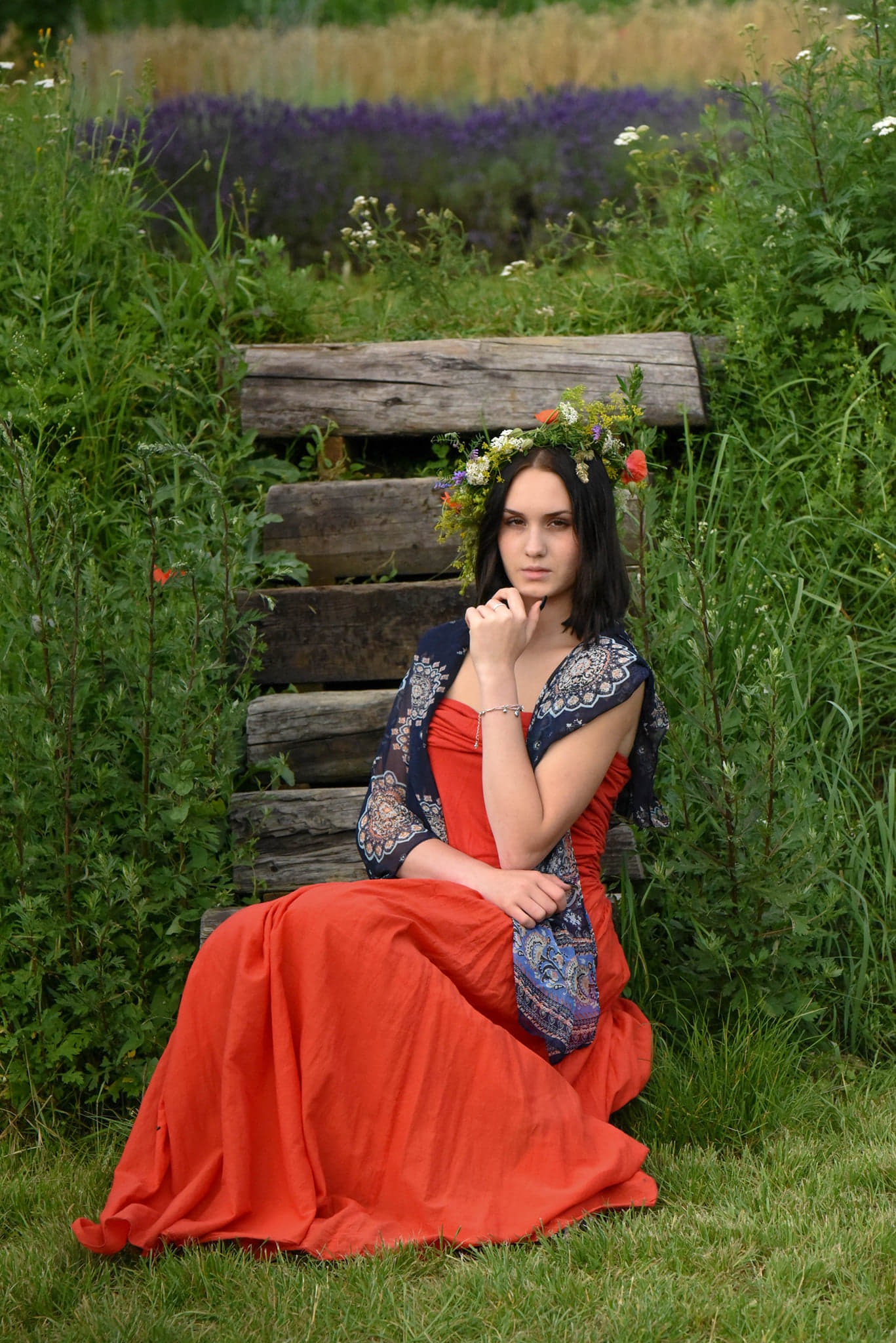 Portret młodej kobiety w pomarańczowej sukni i wianku z polnych kwiatów siedzącej na drewnianych schodach w otoczeniu zieleni i lawendy.