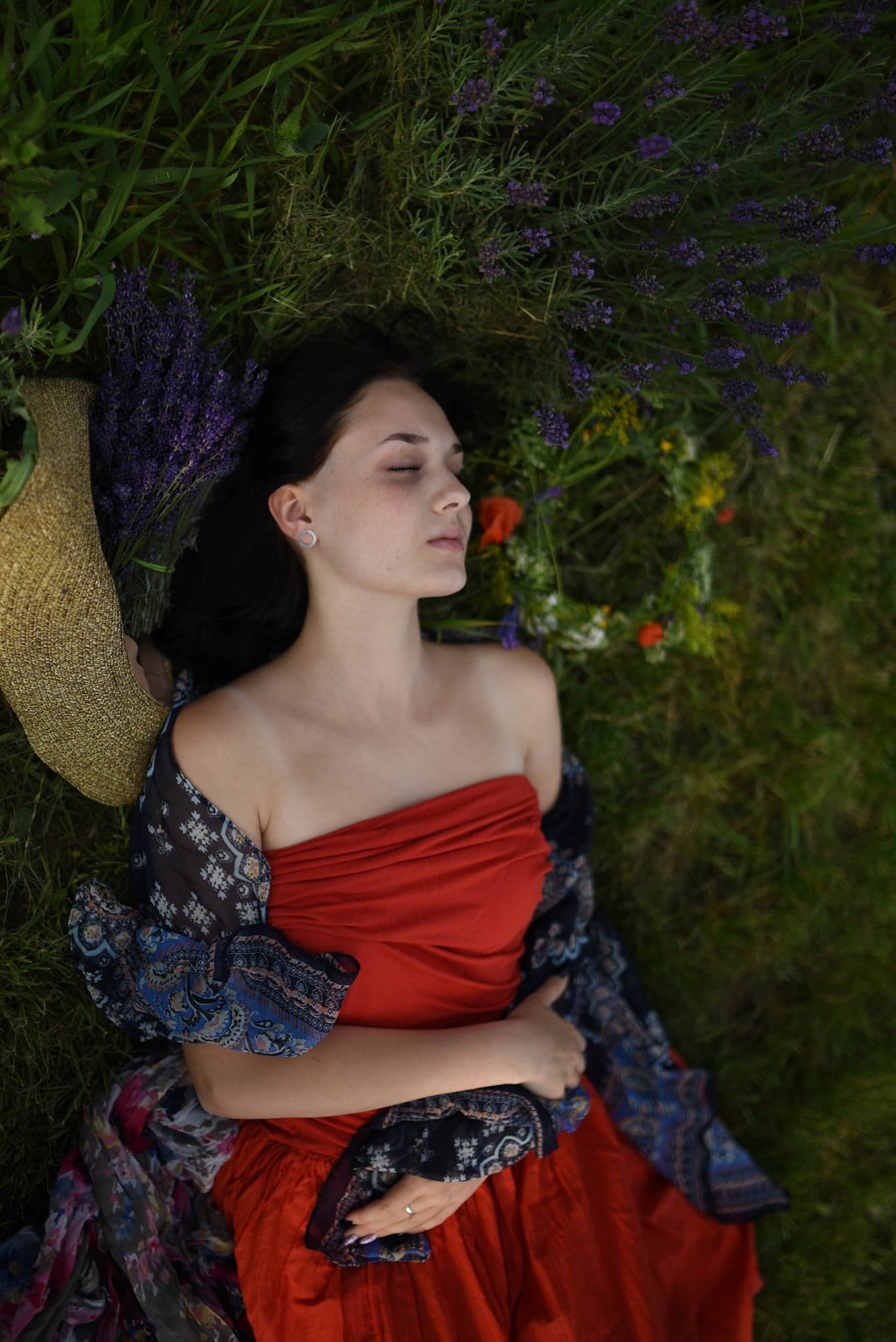 Młoda kobieta w czerwonej sukience leży na trawie z zamkniętymi oczami, otoczona lawendą i kwiatami, z kapeluszem obok głowy i wzorzystą chustą na ramionach.