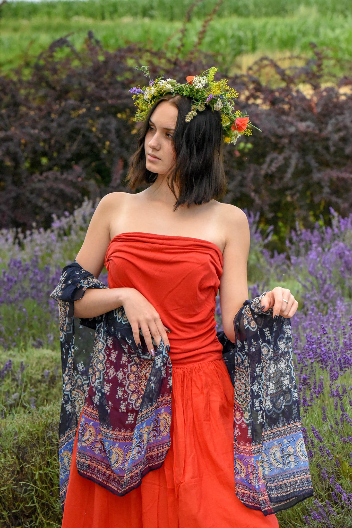 Młoda kobieta w czerwonej sukience i wianku z kwiatów na głowie, pozuje w polu lawendy, trzymając wzorzystą chustę w dłoniach. Uwagę zwracają ciemne paznokcie i pierścionek na palcu.