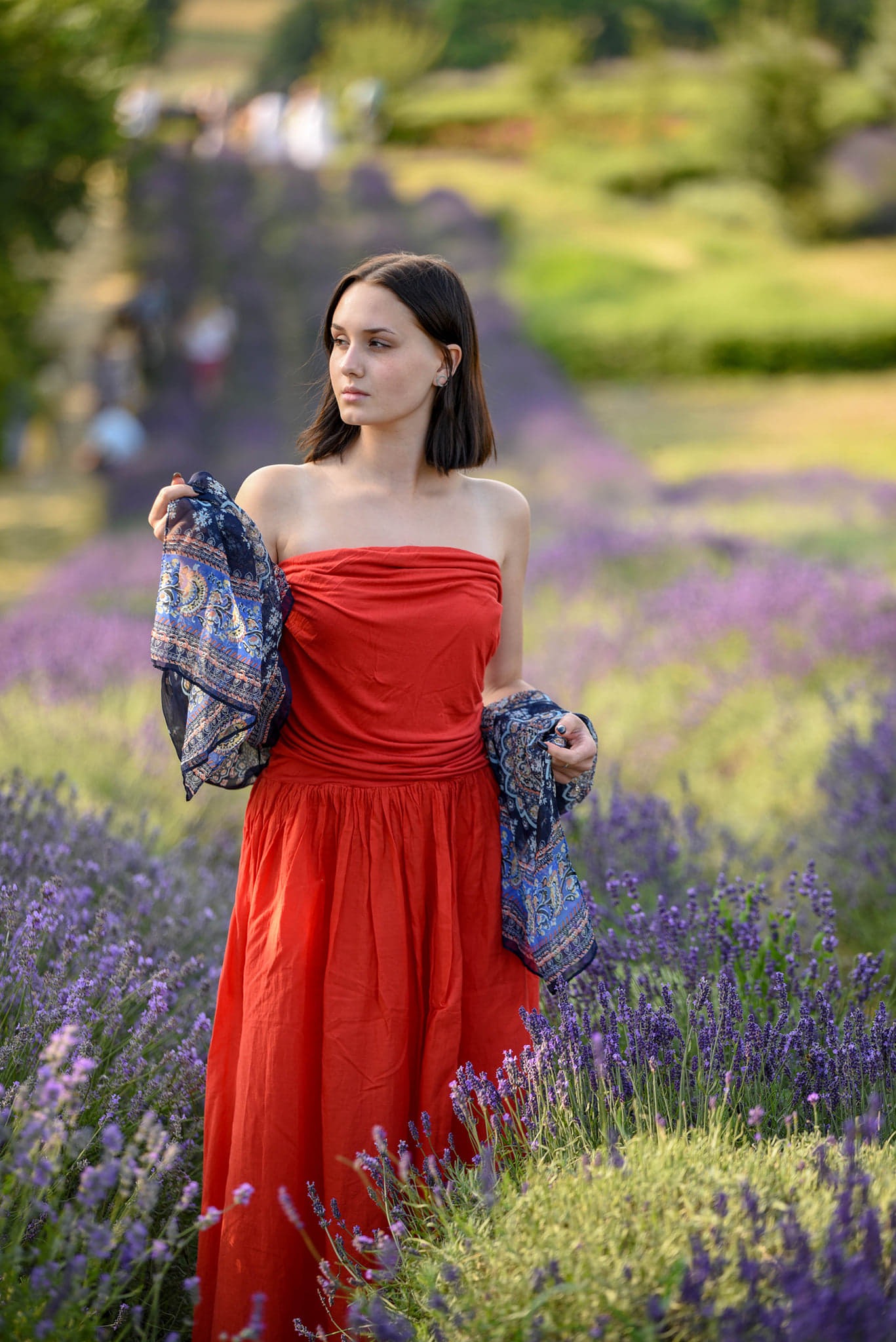 Młoda kobieta w czerwonej sukience i wzorzystej chuście pozuje na polu lawendy, spoglądając w bok.