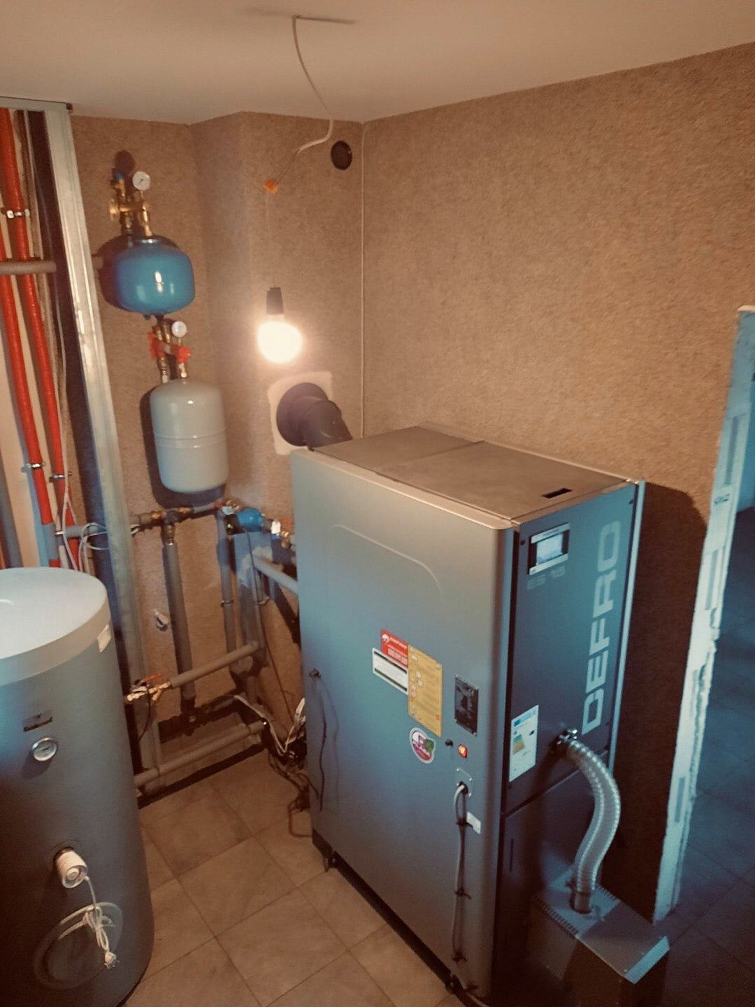 Kotłownia z kotłem na pellet DEFRO, widoczne rury, zbiorniki wyrównawcze, kafelki na podłodze i oświetlenie żarowe.