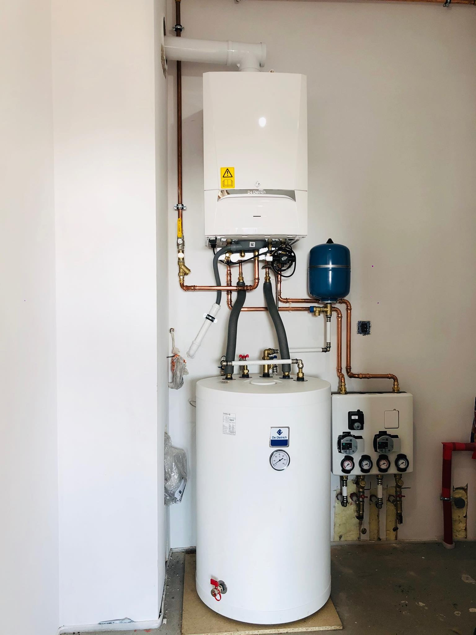 Zestaw kotłowni gazowej z kondensacyjnym kotłem De Dietrich AMC 25kW, emaliowanym zasobnikiem SRK 100L, grupą pompową MT12 na 2 obiegi, widoczne miedziane rury i armatura, całość zamontowana...