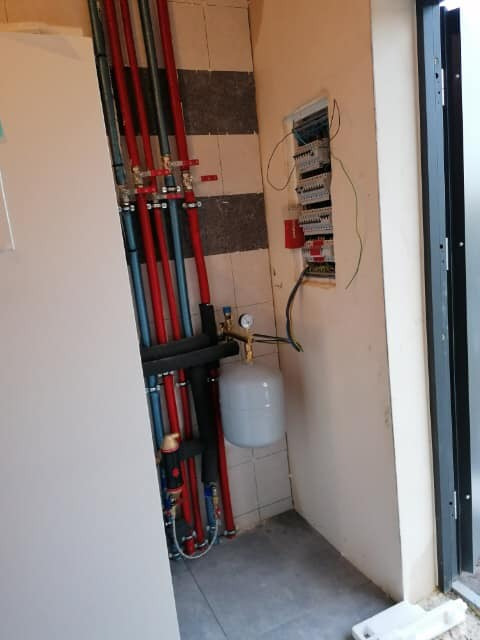 Węzeł instalacyjny z rurami w kolorze czerwonym i niebieskim, zbiornik wyrównawczy z manometrem, oraz skrzynka elektryczna z przewodami na ścianie w pomieszczeniu technicznym.
