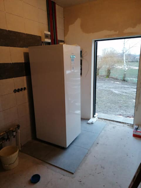 Biała pompa ciepła typu all-in-one firmy Panasonic, z wbudowanym zasobnikiem ciepłej wody, zamontowana na szarych płytkach w surowym pomieszczeniu z widocznymi przewodami instalacyjnymi...