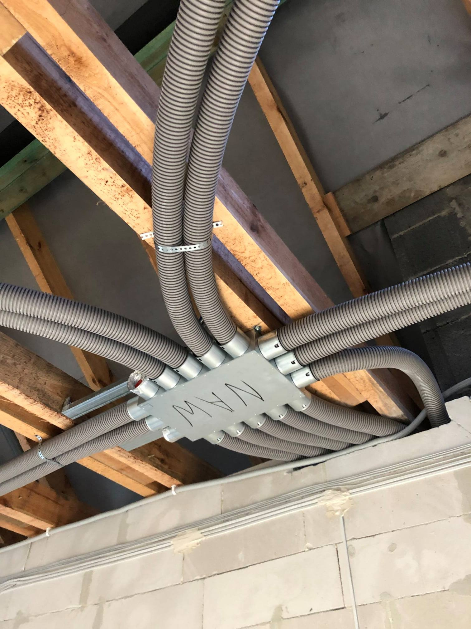 Szare, karbowane rury wentylacyjne rozprowadzone pod drewnianym stropem, połączone metalową skrzynką rozdzielczą z napisem 'MW', widoczne elementy konstrukcji budynku.
