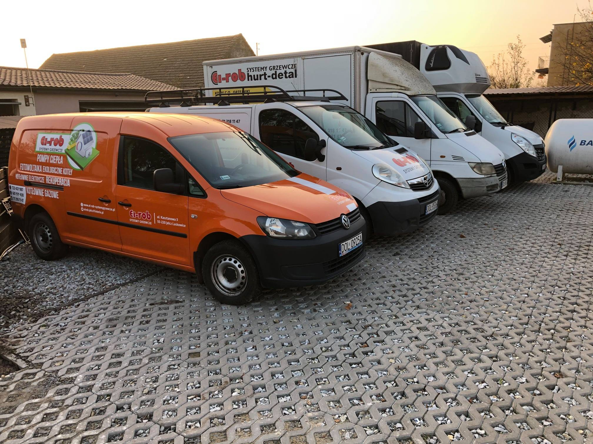 Flota samochodów dostawczych firmy z Jelcz-Laskowic specjalizującej się w systemach grzewczych, w tym pompy ciepła, fotowoltaika, kotły elektryczne i gazowe, zaparkowana na ażurowym parkingu.