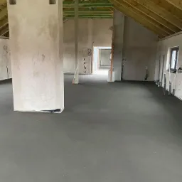 Wnętrze w stanie surowym z wylaną, szarą posadzką jastrychową, widoczne drewniane elementy konstrukcji dachu i kolumna z otworami wentylacyjnymi.