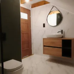 Nowoczesna łazienka na poddaszu z marmurowymi płytkami, drewnianą szafką pod umywalką i okrągłym lustrem zawieszonym na pasku.