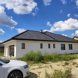 Nowoczesny, parterowy dom jednorodzinny z czarną dachówką, białymi ścianami i brązowymi ramami okiennymi, widziany z perspektywy pod kątem, z zaparkowanym białym samochodem z przodu i budynkiem...