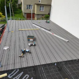 Montaż kompozytowej deski tarasowej na dachu budynku, widoczne narzędzia: wiertarka, poziomica, młotek, puszka z klejem, taśma, czarna membrana z nadrukiem w kratkę. W tle budynek mieszkalny i zieleń.