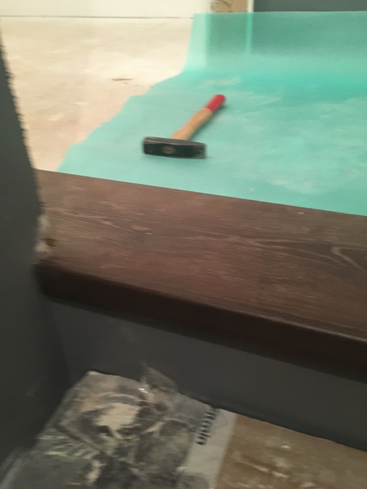 Młotek leżący na zielonej folii ochronnej, widoczny fragment drewnianego progu i opakowanie materiałów budowlanych w dolnej części kadru.