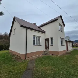 Dwukondygnacyjny dom z beżową elewacją i brązowym dachem, widoczny z perspektywy trawnika, z anteną satelitarną na ścianie i drzewami w tle.