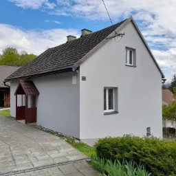 Świeżo otynkowany dom jednorodzinny z brązowym dachem i drewnianą wiatrołapem, widok z boku na tle zieleni i błękitnego nieba z chmurami.