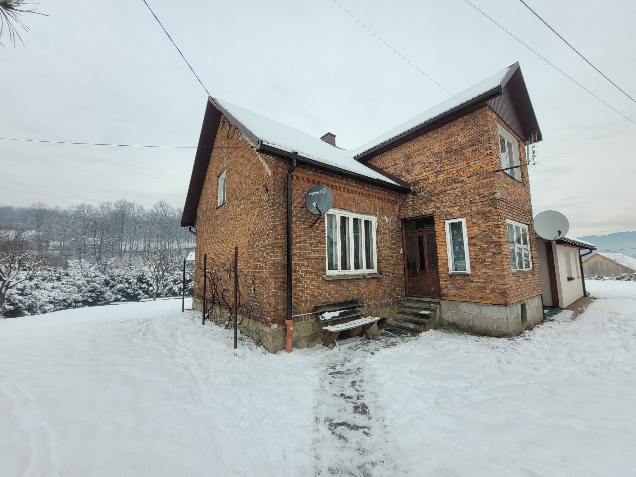 Dwupiętrowy dom z czerwonej cegły w zimowej scenerii. Śnieg pokrywa dach, ziemię i drzewa w tle. Widoczna ławka przed domem i anteny satelitarne na elewacji.