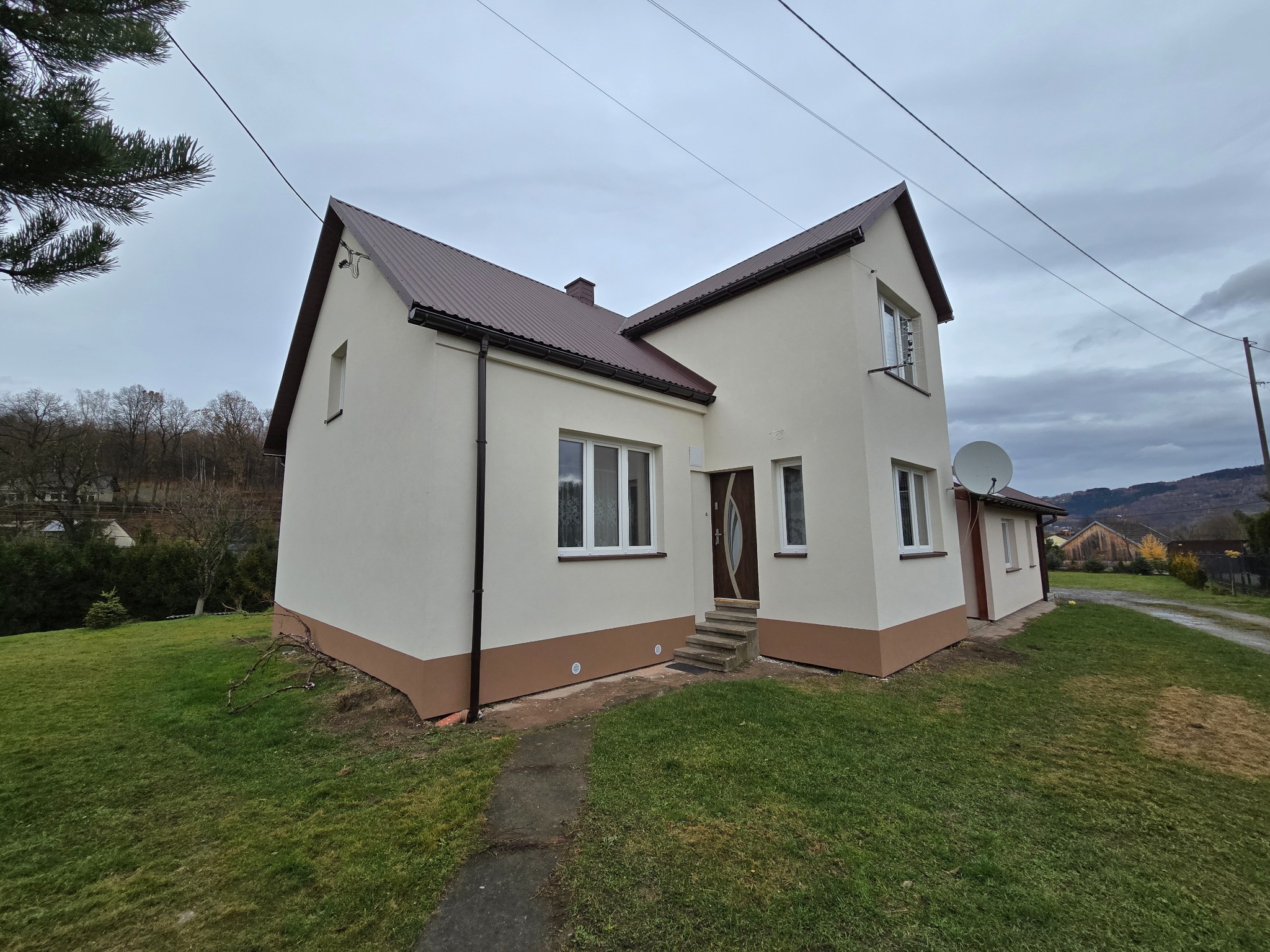 Dwukondygnacyjny dom z beżową elewacją i brązowym dachem, widoczny z perspektywy trawnika, z anteną satelitarną na ścianie i drzewami w tle.