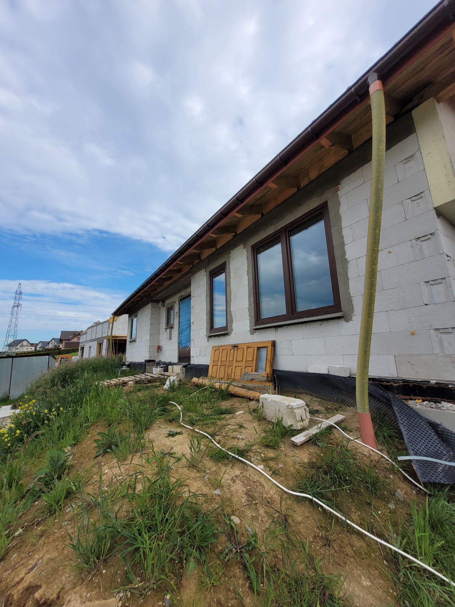 Budowa domu jednorodzinnego z bloczków, widoczne okna z brązowymi ramami i rura wentylacyjna na elewacji. W tle słup wysokiego napięcia i fragmenty zabudowy.