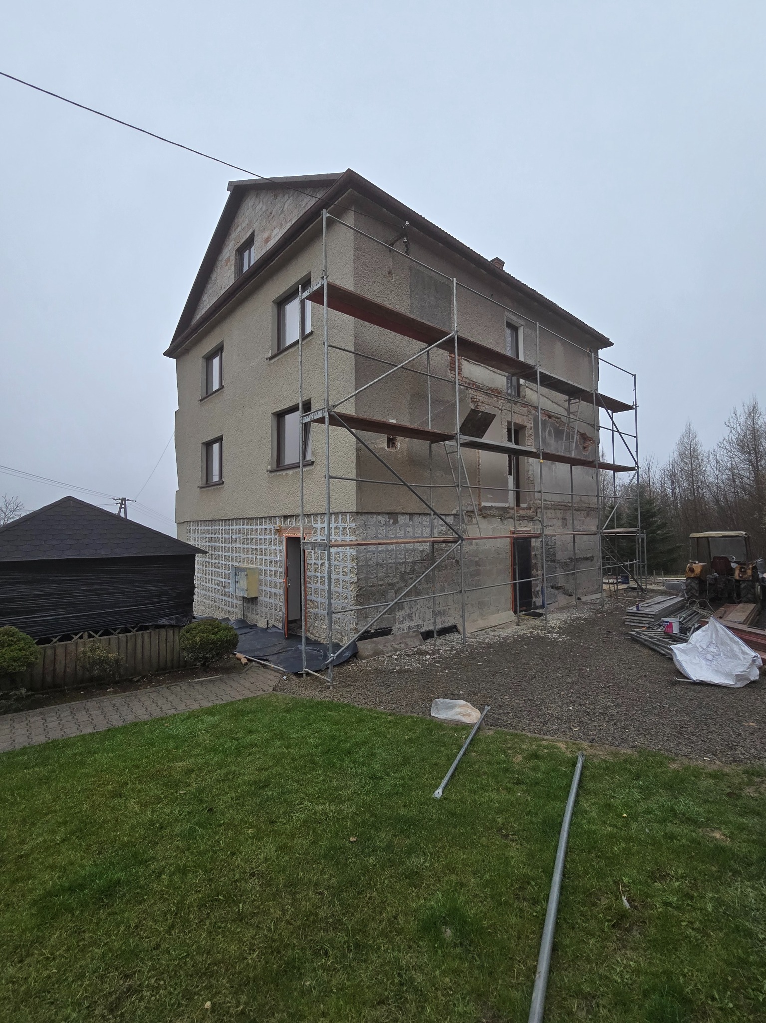 Dwupiętrowy dom w trakcie renowacji elewacji, z widocznymi rusztowaniami i fragmentami starego tynku. Na pierwszym planie trawnik i fragment budynku gospodarczego.