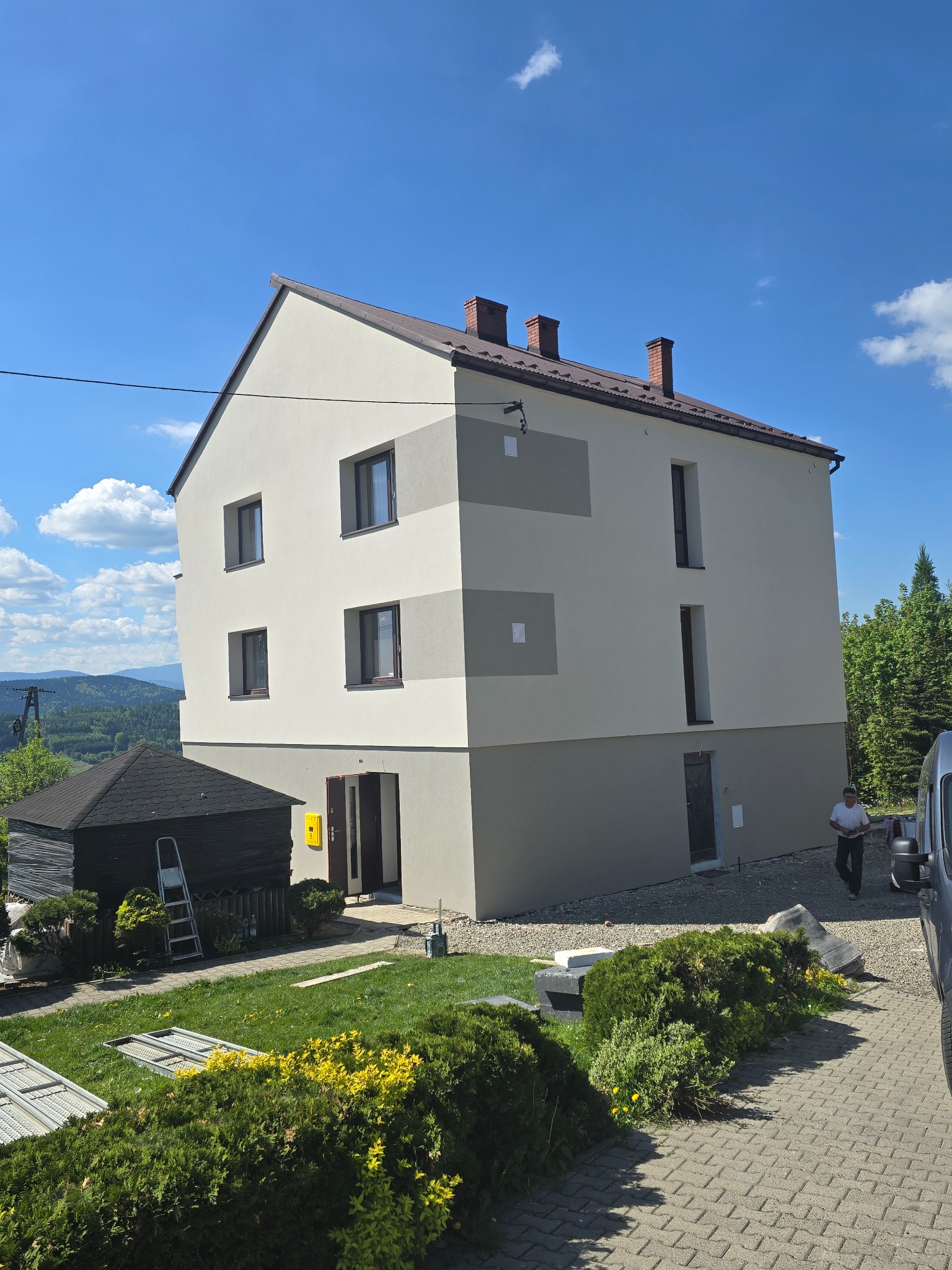 Dwupiętrowy dom po remoncie elewacji z szarymi pasami, na tle zielonych wzgórz i błękitnego nieba. Widoczny fragment chodnika z kostki i pracownik w tle.