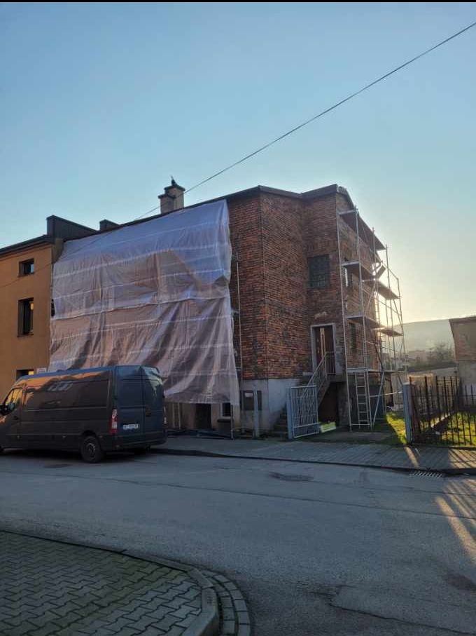 Budynek z cegły w trakcie prac elewacyjnych, osłonięty folią i otoczony rusztowaniem. Na pierwszym planie zaparkowany van.