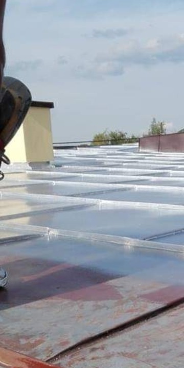 Pracownik w butach roboczych na metalowym dachu podczas mycia, widoczne refleksy wody i budynki w tle.